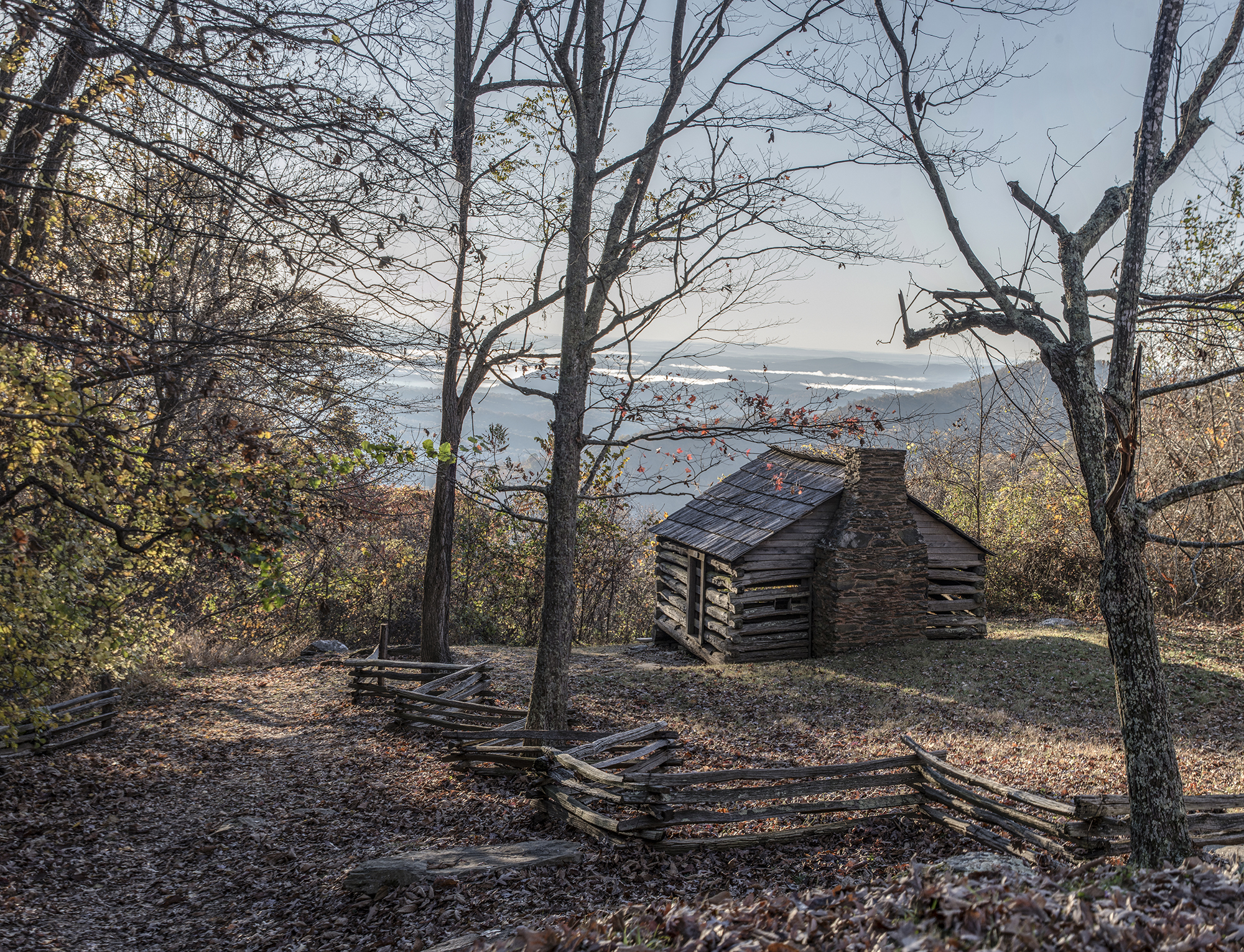 Image of an old, log lookout cabin with zig zag plank stacked fencing surrounding it, perspective from behing the house looking through the trees to a beautiful mountain view
