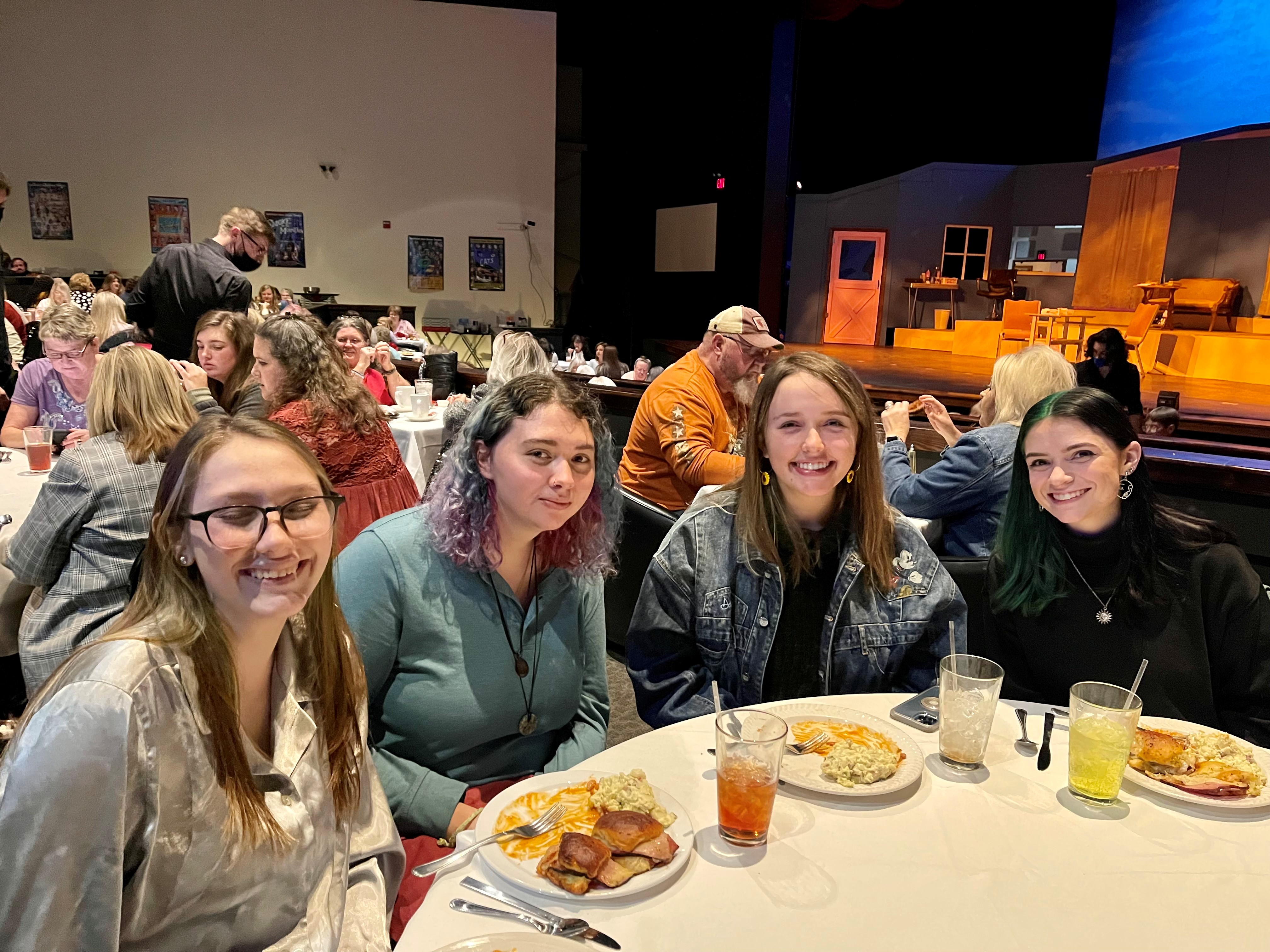 group of four students at a round dinner table sat in front of a theatre stage for a play