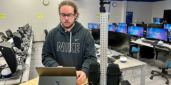 A student works on a laptop with a lab behind him